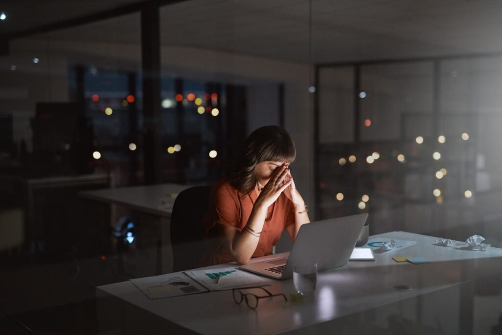 Woman at work desk, late at night with head in hands